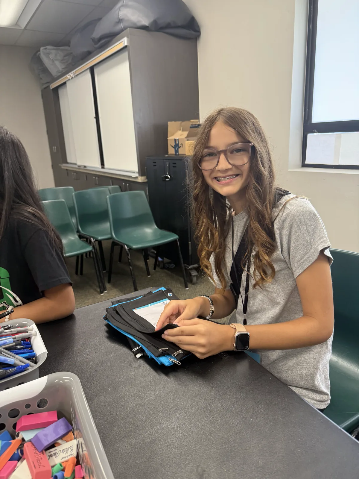 A smiling student sits at a classroom table with a binder and pencil case during a poster-making session
